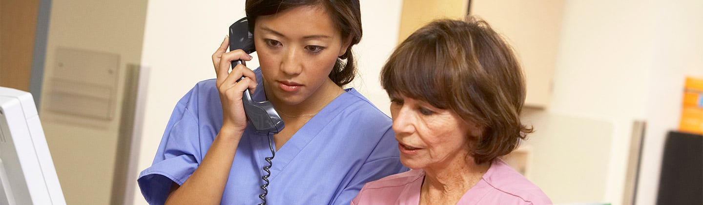 Two nursing professionals wearing scrub outfits are at a desk area in a hospital. One nurse is standing and talking on the phone. The other nurse is seated and holding a folder. Both nurses are looking at information in the folder.