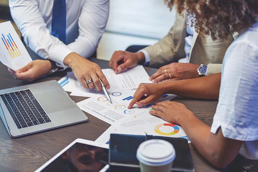 Three people are seated at a table and reviewing financial data spread out before them. Only their arms and hands are visible as they point to sections of a graphic. 