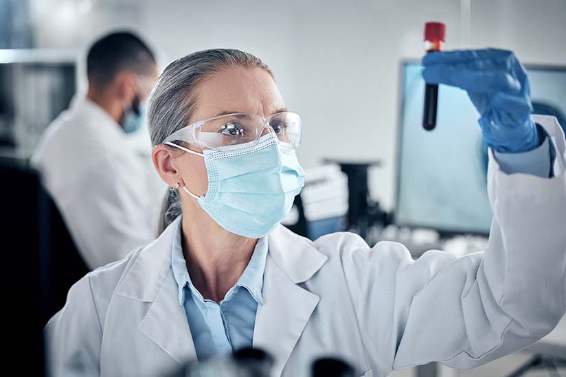 A female nurse is wearing a lab coat, protective eyewear, mask and gloves, and she is holding up and inspecting a blood specimen in a tube.