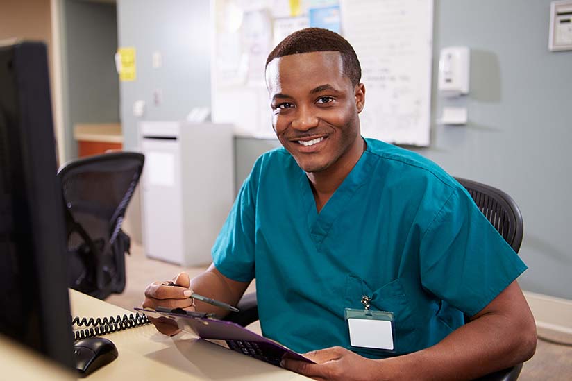 A smiling African American male nurse is seated at a computer workstation in a hospital ward. He is wearing blue scrubs and holding a clipboard and pen. 