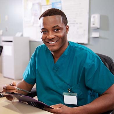 A%20smiling%20African%20American%20male%20nurse%20is%20seated%20at%20a%20computer%20workstation%20in%20a%20hospital%20ward.%20He%20is%20wearing%20blue%20scrubs%20and%20holding%20a%20clipboard%20and%20pen.%20