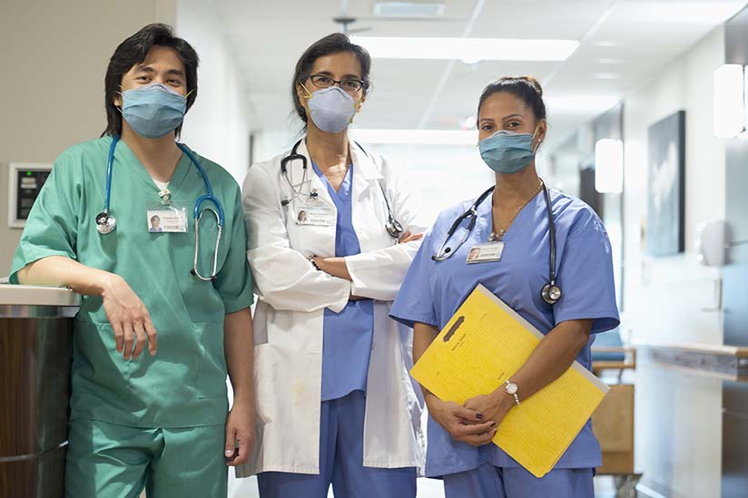 A diverse team of nurses gathers around a table for a meeting, with one standing and leading the discussion, illustrating collaboration and engagement in a clinical setting. 