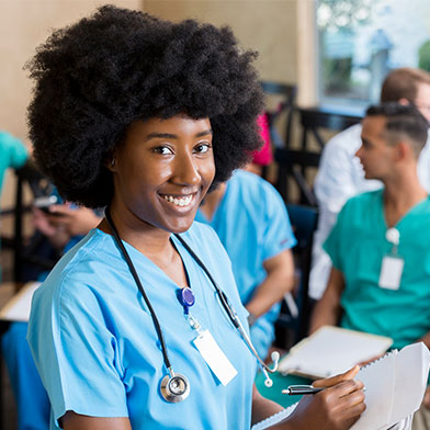 Smiling%20young%20nurse%20with%20an%20afro%20holds%20a%20clipboard%20in%20a%20classroom%20with%20fellow%20nurses%20chatting%20behind.