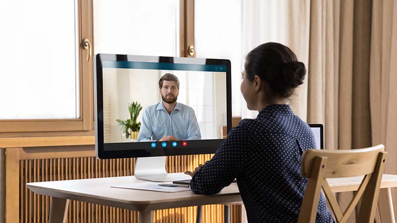 A female nurse is seated at a desk at home and is facing a computer screen. She is speaking with a male interviewer using an online platform.