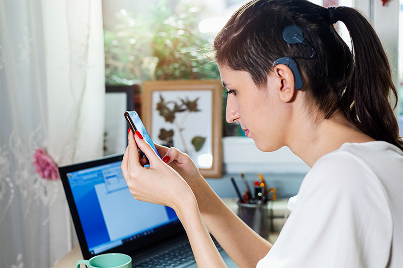 Beautiful young female nurse manager with a visible external cochlear implant is seated at her office desk, and looking at information on her handheld tablet.