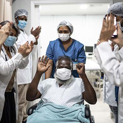 Elderly male patient is being wheeled out of the hospital and hospital staff have lined the hallway and are clapping in happiness for his recovery.