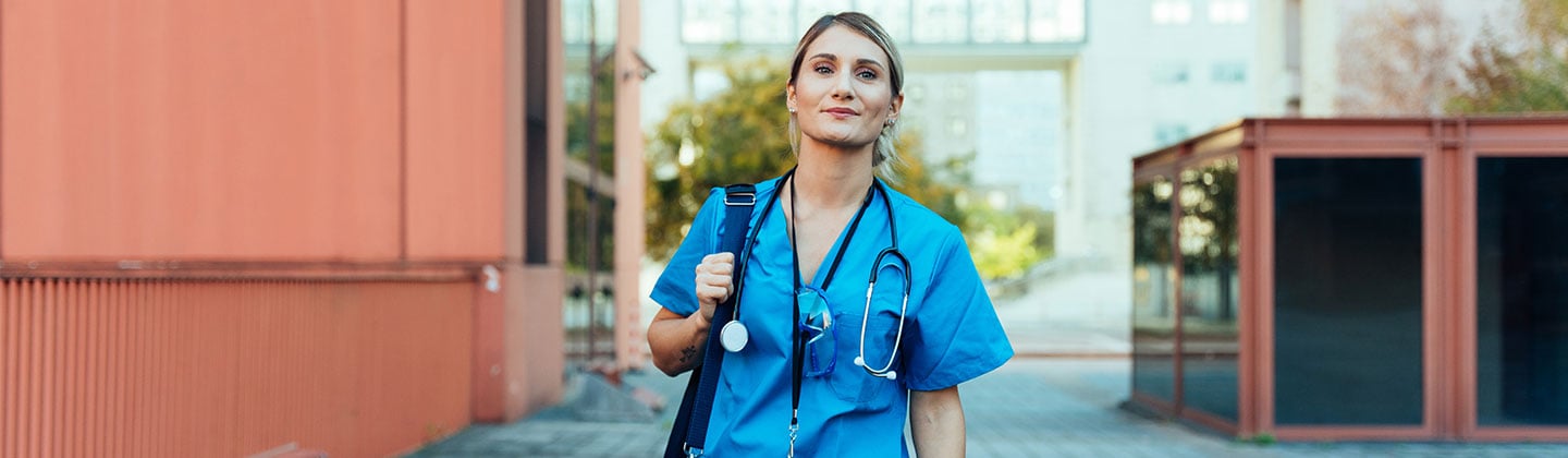 A confident and smiling female nurse wearing blue scrubs is walking outside a hospital on her way to work. She is carrying a blue duffle bag with all the things she will need for a long shift at work.  