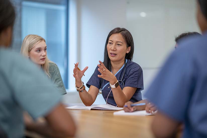 A female nursing leader is seated at a table and meeting with her staff, who are also seated. She is using her hands to emphasize a point, and her staff is looking at her and listening intently.