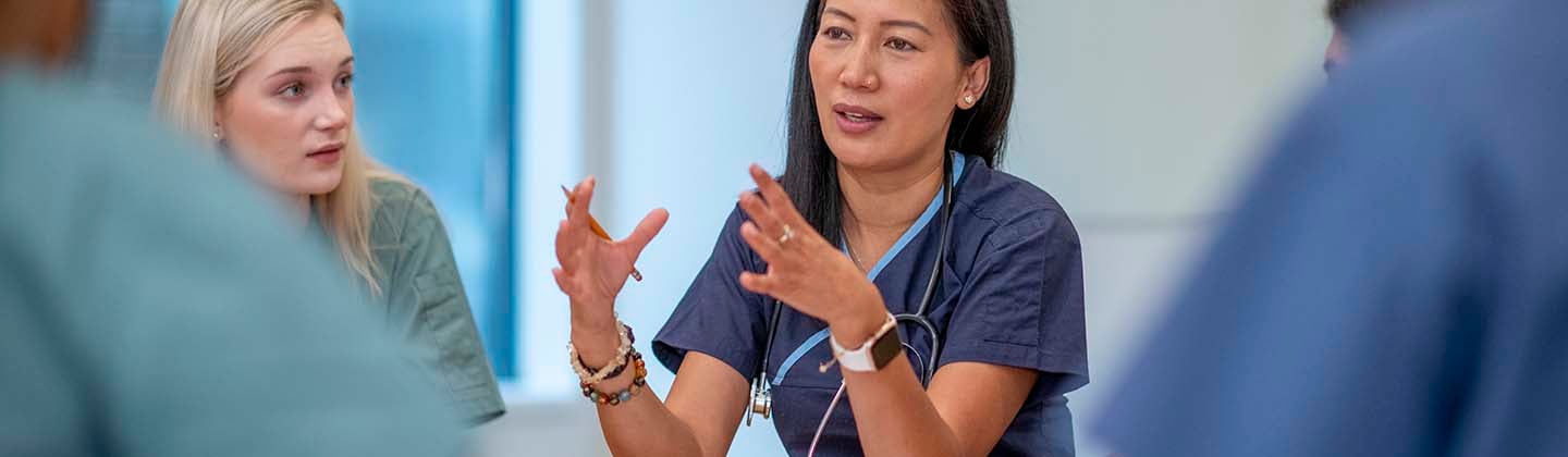 A female nursing leader is seated at a table and meeting with her staff, who are also seated. She is using her hands to emphasize a point, and her staff is looking at her and listening intently.